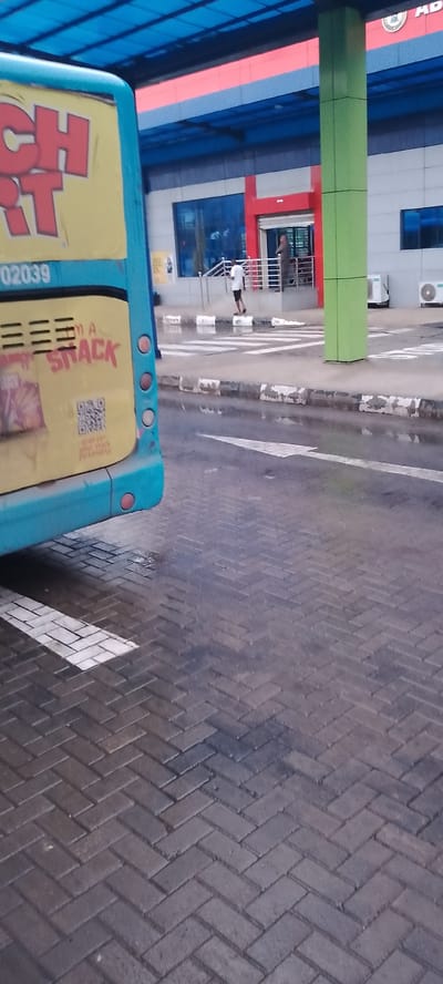 The image shows a street scene in Jibowu, Nigeria. A bright turquoise bus, with yellow and red lettering that reads "CHART" and "SNACK", occupies the left side of the frame. The bus has several circular red lights on its side. In the background, a modern building with blue and red accents and a green pillar is visible. People are seen walking on the sidewalk in front of the building. The ground is wet, suggesting it has recently rained, and the sky is overcast. A white arrow is painted on the wet road, indicating a direction. There is also a QR code on the side of the bus. The overall impression is of a busy, wet day in an urban Nigerian setting.