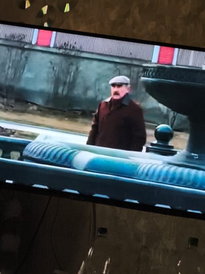 The image shows a man wearing a flat cap and a dark coat, standing in what appears to be an outdoor setting. He has a prominent mustache. In the foreground, there is a railing with a blue and white striped pattern. The background is out of focus but suggests a building with red and white elements. The overall scene has a muted color palette. No specific details regarding the city of Monino, Russia, can be deduced from the image.