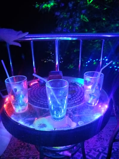 The image shows three drinking glasses, each with a straw, positioned on a round table with a mosaic-patterned top. The table is illuminated with blue and red lights, creating a neon effect. A railing with vertical supports is visible in the background, along with some green foliage. A hand rests on the railing, and there is a hint of patterned carpet below the table. Based on the location (Moo 4, Thailand), it is likely an outdoor bar or lounge area.