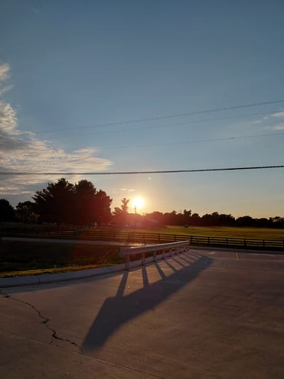 The image depicts a sunset scene in Paint Lick, United States. The sun is low in the sky, casting a warm glow over a rural landscape. A paved road or parking lot occupies the foreground, with a concrete barrier and a long shadow stretching across the surface. Beyond the road, a grassy field is bordered by a wooden fence. In the middle ground, a cluster of trees is silhouetted against the bright sky. The sky itself is a gradient of blue and orange, with a few wispy clouds. Power lines are visible in the upper portion of the frame. The overall atmosphere is serene and peaceful.