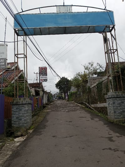 The scene depicts an upward-sloping paved residential street in Melakudikadu, India, under an overcast sky. A large, weathered metal archway, featuring a blank blue rectangular panel, spans the road in the foreground. This archway is supported by rusty square-section metal framework posts, each resting on a stone-patterned masonry pillar. Numerous black utility wires crisscross overhead, attached to the archway structure and extending down the street.
To the left of the road, a low purple fence is visible, behind which are residential structures with terracotta-tiled roofs. A white rectangular sign on a pole, partially obscured, displays text including "KEB", "ARI AM", and "Cianjur". Further down the street on the left, another prominent black sign with white text reads "TOKO MILA" alongside other script.
To the right of the road, beyond the archway's support pillar, there is a stone retaining wall partially covered in green vines and sparse vegetation. This wall separates the road from higher ground where additional residential structures are visible. The asphalt road surface appears worn and uneven, with various patches and stains. No individuals are present, and no actions or interactions are occurring. The environment suggests a built-up area with a mix of natural elements and infrastructure.