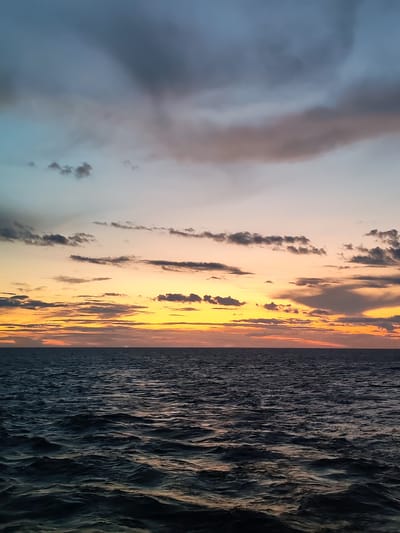This is a photograph of a sunset over the Rybinsk Reservoir, part of the city of Mologa, Russia. The horizon is marked by the calm, dark water of the reservoir, with a few distant boats visible as small dark shapes. Above the water, the sky displays a gradient of colors, transitioning from the deep oranges and yellows of the sunset near the horizon to softer pinks, blues, and grays further up. Wispy clouds are scattered across the sky, some catching the last rays of sunlight and appearing in shades of pink and orange, while others are darker gray. The overall impression is one of peaceful twilight over a large body of water.