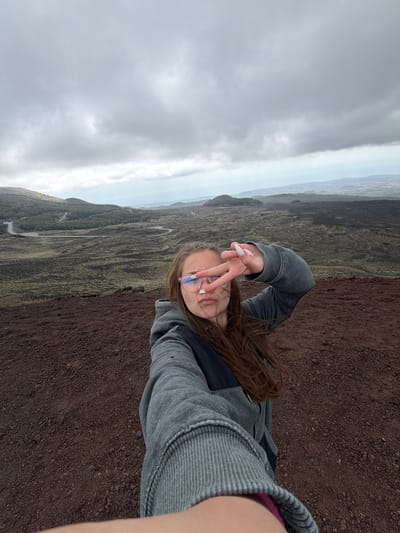 A person stands in a landscape near Nicolosi, Italy, holding up two fingers in a peace sign towards the camera. The background shows a winding road and a landscape with sparse vegetation. The sky is cloudy. The ground is covered in dark, granular material. The person is wearing glasses and a grey jacket.