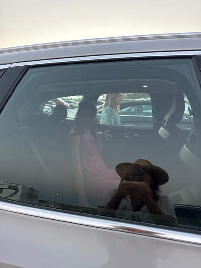 The image displays a multi-layered scene captured through the rear passenger window of a light-colored vehicle, situated in an outdoor parking area in Almancil, Portugal.

In the foreground, a prominent reflection on the tinted window shows a person from the chest up, wearing a wide-brimmed straw hat and a light-colored top. This individual is holding a smartphone with both hands, pointing it towards the vehicle's interior, indicating they are the photographer. A ring is visible on their left hand.

Inside the vehicle, a young female with dark, curly hair, dressed in a pink sleeveless garment, is seated in the rear passenger compartment. She is looking directly towards the camera with a wide smile, and her right hand is raised with fingers splayed in a gesture. The vehicle's interior features dark upholstery and headrests, with a light-colored roof lining visible.

Through the vehicle's windows, in the background, an outdoor parking lot is visible. Another female individual with light-colored hair, wearing a patterned light-blue and white top, is observed walking from left to right in the mid-distance. Numerous other parked cars of various colors populate the background. The sky is a uniform light grey, suggesting overcast conditions or soft lighting. A light pole is faintly visible in the background, its reflection also present on the vehicle window. The vehicle itself has chrome trim detailing along its window lines.