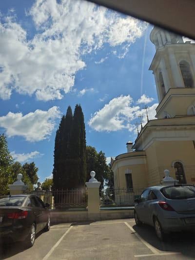 The image shows a view of a church building in Chekhov, Russia. The church is light yellow with white architectural details and a bell tower. In front of the church, there is a paved parking lot with two cars. A dark sedan is parked on the left, and a light blue hatchback is parked on the right. The sky is blue with scattered white clouds. Tall, dark green cypress trees are visible in the middle ground, and lush green trees and bushes surround the church. A wrought-iron fence with decorative posts separates the parking lot from the church grounds. The sun is shining, creating highlights and shadows.