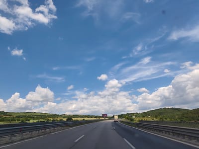 A highway with a guardrail runs through a rural landscape under a blue sky with white clouds. In the distance, a truck is driving on the highway, and a billboard is visible on the side of the road. Hills covered with trees and vegetation can be seen in the background. The location is the Eleshnitsa, Bulgaria area.