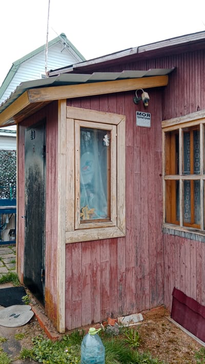 A close-up captures the corner of a weathered, red-painted wooden outbuilding in what appears to be a rural or suburban setting. A green-roofed structure peeks out in the background, suggesting a residential area. The outbuilding has two windows with worn wooden frames, and a black door with the number "40" painted on it. The window to the left contains sheer curtains with a few decorative items visible inside. A surveillance camera is mounted above a small sign with writing in Russian that translates to "My Territory". A large bottle of water sits on the ground near the corner of the building. The ground is covered with patchy grass and weeds. The scene is located in the city of Былина Снт, Russia.