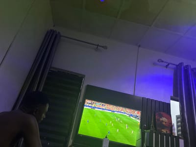 In a dimly lit room, a young man sits facing a television screen displaying a soccer match. The room features a dark, slatted wall behind the television, adorned with a small Eiffel Tower replica and a framed picture of a sunset over a palm tree landscape. Blue and green lighting illuminates the scene, casting shadows on the walls and ceiling, which are paneled with acoustic tiles. Curtains hang on either side of the room, and a large window is visible on the right. The man, partially visible from the back, is shirtless. On the television screen, a soccer game is in progress, with players in blue and white uniforms on a green field. A score graphic at the top of the screen indicates "90:00" and "4:2". The setting appears to be a personal living space, possibly in Naraguta Hausawa, Nigeria, given the context of the request.