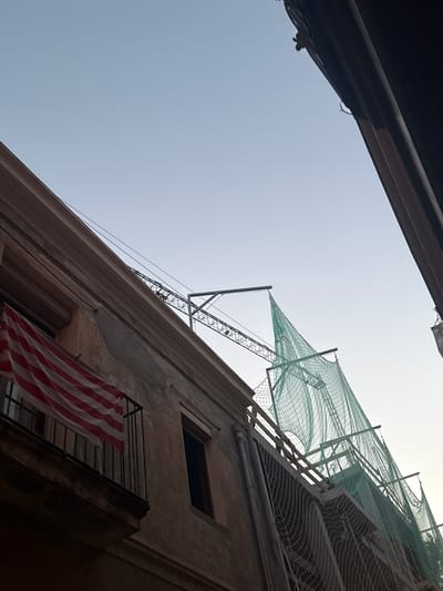 The image shows a close-up, low-angle shot of a building under construction or renovation. A balcony on the left side of the frame displays a flag with red and white horizontal stripes, possibly the flag of the Basque Country or a similar regional flag. Above the building, a construction crane is visible, along with green safety netting stretched across the scaffolding. The sky is clear and a pale blue, suggesting it is either early morning or late afternoon. The architecture of the building appears to be old, with weathered plaster. The scene is set in an urban environment, implied by the presence of buildings and construction. There are no people visible in the image. The location is identified as Sant Boi de Llobregat, Spain.