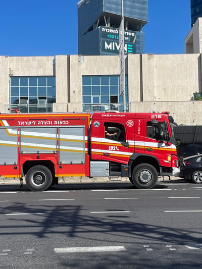 A red fire truck with white and yellow stripes is parked on a city street in Kfar Saba, Israel. The truck has the Hebrew text "כבאות והצלה לישראל" (Fire and Rescue Services Israel) written on its side. The truck also displays the number "102" and a website address "www.102.gov.il". In the background, there is a modern building with large windows and signs. A black car is visible to the right of the fire truck. The street has white lane markings and shadows of trees or structures are cast on it. The sky is clear and blue.