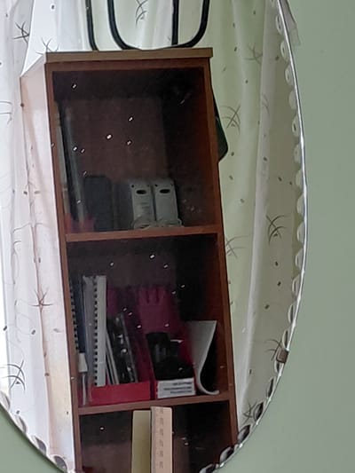 The image captures a reflection in a decorative, oval-shaped mirror with a scalloped, etched edge. The mirror is positioned against a plain, light green wall.
The reflection prominently features a wooden multi-compartment shelf or box. The shelf is divided into at least three visible sections, each containing various items. In the leftmost compartment, two light-colored, possibly metal, objects with oval cutouts are stacked, with other darker items below them. The central compartment contains a red-lined container holding several spiral-bound notebooks or binders and a white rectangular object with faint, undecipherable text, possibly "INFORMATION" or "ILLUSTRATIONS." The rightmost compartment holds a light wooden ruler with faint measurement markings and a stack of paper or a book. Small white specks, likely reflections of light sources, are visible within the compartments.
Above the wooden shelf in the reflection, a light-colored, possibly white or light green, curtain or fabric is visible. This fabric is patterned with abstract, dark, calligraphic-like lines and scattered small dots or squares.
No people are present, and no activity is taking place. The scene suggests an indoor setting, likely a study or office area in Nadym, Russia. The lighting is ambient, and no specific time of day can be determined.