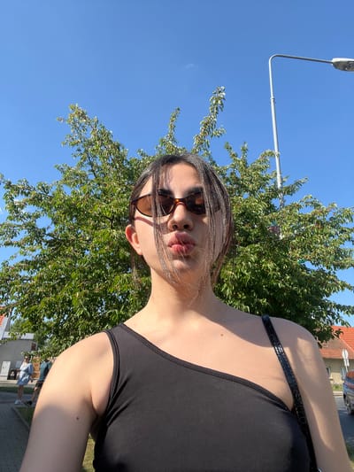 An outdoor, low-angle, medium shot captures a young woman with dark hair wearing sunglasses and a black one-shoulder top, posing with pursed lips for a selfie. Behind her, a lush green tree with small red fruits fills the background, partially obscuring a bright blue sky. To the right, a tall gray streetlight stands prominently. In the distance, a street and buildings with red roofs are visible, with two people walking on a paved path. The scene appears to be on a sunny day, possibly in Dobřany, Czechia, given the architectural style and vegetation.