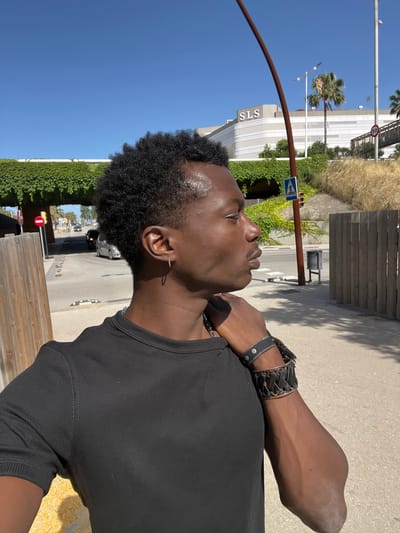 A young Black man is pictured in a medium shot, facing right, with his head turned slightly towards the camera. He has short, curly black hair and is wearing a black t-shirt. His left hand rests on his right shoulder, and he has two bracelets on his right wrist. The background suggests an outdoor urban environment with a clear blue sky. Behind him, a building with "SLS" signage is visible, along with palm trees and dry vegetation. A pedestrian crossing sign and a speed limit sign (50) are also present. A wooden fence runs along the right side of the frame. The man appears to be outdoors on a sunny day. This scene is located in Sant Adrià de Besòs, Spain.
