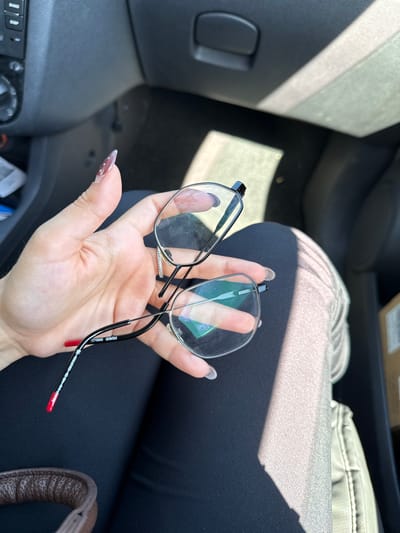 An interior view of a vehicle in Bruchköbel, Germany, shows a person's left hand holding a pair of black-framed Hugo 5005 prescription eyeglasses. The hand displays manicured nails, with one visible nail featuring white polka dots on a reddish-brown polish and another having solid red polish on its tip. A silver-toned ring adorned with small white stones is present on the ring finger. The eyeglasses feature clear, somewhat smudged lenses, and the temple arm clearly displays the branding "HUGO 5005."
The individual is wearing dark clothing on the left leg, while the right leg or adjacent car seat appears covered in a lighter grey, textured fabric. Bright sunlight streams into the vehicle, creating distinct bright streaks and shadows across the interior, including the person's leg and the car's headliner. Visible car interior elements on the left include a dark dashboard section with audio controls labeled "BAND" and "DISP," along with a rotary knob. A dark overhead compartment handle is visible on the upper right side of the frame. In the lower left, a brown, textured strap, likely from a bag, is partially visible. A cardboard box is partially visible in the bottom right corner of the frame, suggesting its presence inside or directly outside the vehicle.