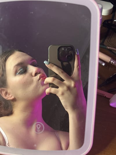 A close-up, mirrored selfie shows a person, likely female, with their head tilted and lips pursed. They are wearing makeup, including blue eyeliner and what appears to be lip gloss. Their right hand is holding a smartphone, obscuring part of their face and chest. The background is dark, with a pink and purple light illuminating the person's neck and chest. The lighting suggests an indoor setting, possibly a bedroom or vanity area. There are makeup brushes and other beauty products visible on a wooden surface to the right of the frame. No specific location in Bishkek, Kyrgyzstan can be determined from the image.