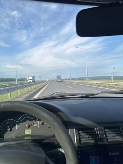 This media file captures a first-person perspective from the driver's seat of a car traveling on a multi-lane highway, likely near Усть-Сыны, Russia. The view looks forward through the windshield on a bright, clear day, with a blue sky sparsely dotted with wispy white clouds.

Inside the car, parts of the dashboard and steering wheel are visible, primarily on the right side of the frame. The speedometer indicates a speed of approximately 100 km/h, with "km/h" clearly legible. A digital display below the speedometer shows "190902" (likely the total odometer reading) and "1826" (possibly a trip meter). A tachometer marked "x100min-1" is also present. The dashboard features air vents, a hazard warning button with a red triangle symbol, and part of an infotainment system displaying "Bluetooth". A rearview mirror is partially visible in the upper left corner.

Outside, the highway stretches ahead with multiple lanes, bordered by guardrails and grassy verges. Utility poles or lamp posts line the road. Several other vehicles, including a white truck, are visible in the distance. The scene depicts a routine journey on an open road under good weather conditions.