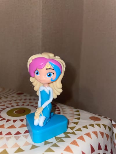 A toy figurine of a young girl with blonde hair streaked with pink and blue stands on a blue base. She is wearing a blue dress with white accents. The figurine is kneeling on a patterned tablecloth. The background is a plain, textured wall. The image is taken indoors, likely in a home. There is no identifiable location in the city of Laishevo, Russia.