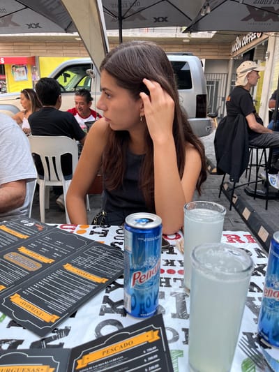 Here is a detailed description of the image:

The photograph captures an outdoor dining scene, likely a restaurant or cafe, in San Francisco Totimehuacán, Mexico. A young woman with long, dark brown hair is seated at a table. She is wearing a black top and has her hand near her face, possibly adjusting her hair.

On the table, there are various items: a blue can of "Peñafiel" mineral water, two tall glasses with a light-colored beverage (possibly lemonade), and a menu with sections labeled "Burguesas", "Pescado", "Carnes", "Entradas", "Alitas", and "Postres" (Burgers, Fish, Meats, Entrees, Wings, and Desserts).

Other people are visible in the background at adjacent tables. A man is wearing a white shirt with a red and white jacket. Another man is wearing a black shirt and is seated at a table. There is a portion of a white plastic chair also visible. The background shows a glimpse of the town, with a white delivery truck and buildings.

An umbrella provides shade above the table.