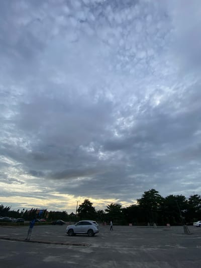 The image shows a cloudy sky above what appears to be a parking lot. The scene is likely at dusk or dawn, given the lighting. The parking lot is sparsely populated with cars. Trees and a silhouette of buildings are visible in the background. One person stands near parked cars in the foreground. The location is possibly Sangotedo, Nigeria, based on the request.