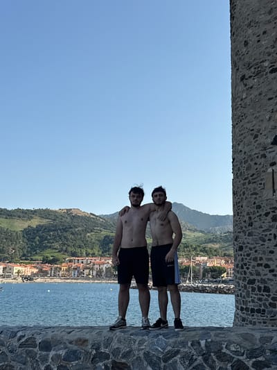 This media file depicts two shirtless men standing on a stone wall overlooking a picturesque coastal town in Collioure, France. The man in front, with dark hair, is positioned slightly lower and to the left, with his arm around the shoulders of the man behind him. The second man, also with dark hair, is standing behind and slightly above the first. Both men are wearing dark shorts. The man in front is wearing blue and white shorts, while the man behind him is wearing black shorts. They both appear to be relaxed and in good spirits.

The background reveals a vibrant scene. The town, with its terracotta roofs and colorful buildings, cascades down a hillside towards the sparkling blue sea. Lush green vegetation covers the slopes of the mountains behind the town. The sea is calm, with a few small boats visible in the distance. The sky is a clear, bright blue, suggesting a sunny day. The stone wall they are standing on is textured and appears to be part of an ancient structure, possibly a fortification or a terrace. The image is taken from a low angle, looking upwards towards the men and the town.

The overall impression is one of a serene and idyllic vacation scene. The time of day appears to be mid-day, given the bright sunlight and clear sky. The location cues strongly suggest Collioure, France, known for its charming Mediterranean atmosphere and artistic heritage. There is no visible text in the image.