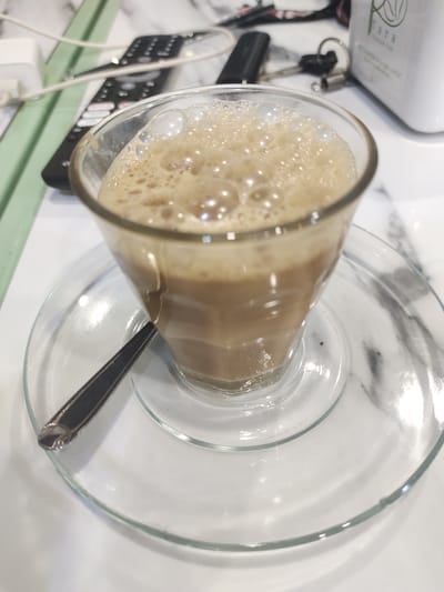 This media file features a close-up shot of a frothy beverage in a clear glass, placed on a clear glass saucer with a dark spoon resting on it. The drink is a light brown color with a thick layer of foam and bubbles on top. The background is slightly blurred, showing a white surface with a marble-like pattern.
In the background, several objects are visible, suggesting a casual setting. To the left, there is a black remote control and a white object, possibly a charger or a power adapter, connected by white wires. To the right, a white container with green text and an image of leaves is partially visible, along with car keys attached to a keyring. The overall impression is of a table at home or a cafe.
The lighting suggests it might be daytime, with the bright light reflecting off the glass surfaces. There are no people visible in the image. The focus is entirely on the beverage and its immediate surroundings. The visible text on the white container appears to be "Jara," with a leaf logo above it. The scene evokes a sense of relaxation and enjoyment of a beverage. The location context is Meunasahaluedrienlhoksukon Barat, Indonesia.