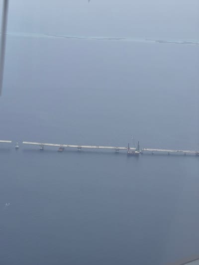 The image shows a long, thin, white structure running vertically through the center of the frame, appearing to be a pier or walkway over water. The structure is composed of multiple segments, some of which are broken or partially submerged. There is construction equipment, including a crane, on one of the intact sections. The water is a uniform gray-blue color. The image is taken from an elevated viewpoint. The left side of the image is obscured by a light gray haze or fog. The location is suggested to be the city of Malé, Maldives.