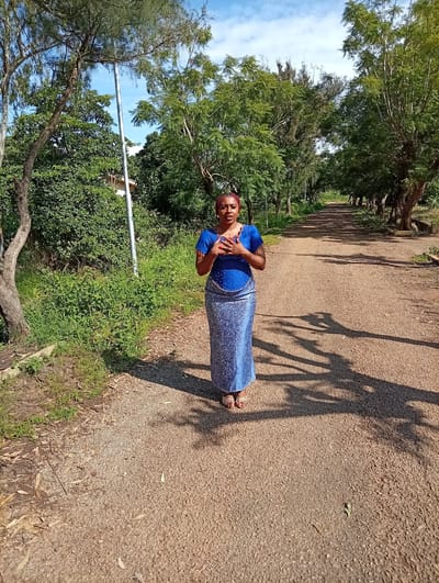 A woman stands in the middle of a gravel road, framed by trees on either side. She is wearing a blue dress with a patterned skirt and a red headscarf. Her hands are clasped at her chest. The road appears to be in a rural or natural setting, with lush green vegetation on the left side of the image and trees lining the right. Shadows of the trees are cast across the road, suggesting it is daytime, likely afternoon, with the sun positioned to the right. The sky above is blue with some scattered clouds. The overall mood is serene and peaceful. There is no visible text in the image. The location is identified as Tambes Mile Four, Nigeria.