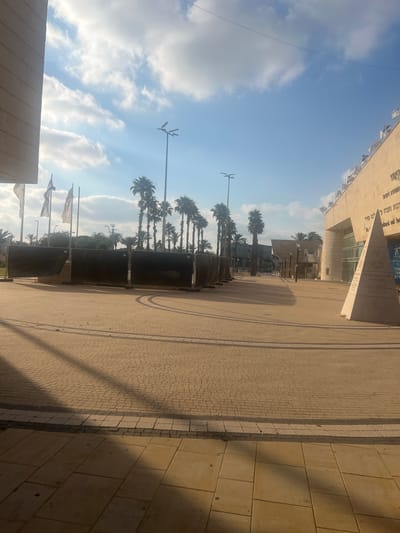 This is a vertical, eye-level, outdoor photograph taken on a sunny day with a clear blue sky and scattered clouds. The scene depicts a modern architectural complex with a paved walkway and a row of palm trees.

In the foreground, a paved area, likely a plaza or promenade, is visible. The paving stones are a light tan color, arranged in a geometric pattern. To the right of the paved area, a section of a building with large, pale stone blocks is visible, casting a long shadow.

A line of tall palm trees with their characteristic fronds stands in the middle ground, separating the paved area from a more distant building. The trees are silhouetted against the sky. Behind the trees, parts of a large, modern building complex are visible. This building features large glass windows and elements of light-colored stone cladding. A triangular monument or architectural feature is prominent in front of this building.

Several flags are visible on poles in the middle ground, though their designs are not clearly discernible. A lamppost with multiple lights is also present. The overall setting appears to be a public space, possibly a cultural center or civic area, given the text visible on the building.

The text on the building, partially visible and in Hebrew, mentions "Or Akiva" (עיר עקיב) and appears to indicate a cultural and social center (מרכז תרבות ותחברה). Some English text is also visible, including "SOCIAL CENTRE."

The sunlight suggests it is daytime, likely late morning or afternoon, given the angle of the shadows. The weather appears pleasant and warm. No people are clearly visible in the image.