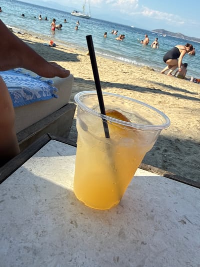The image captures a sunny day at a beach in Thassos, Greece. In the foreground, a plastic cup filled with a yellowish beverage and ice, with a slice of orange and a black straw, sits on a speckled white table. To the left, a person's leg and foot, with sand on them, rest on a blue and white striped cushion. The background shows a sandy beach and the clear blue sea. Several people are visible in the water, swimming and enjoying themselves. A sailboat is anchored in the distance. The overall scene depicts a relaxed atmosphere at a seaside resort.