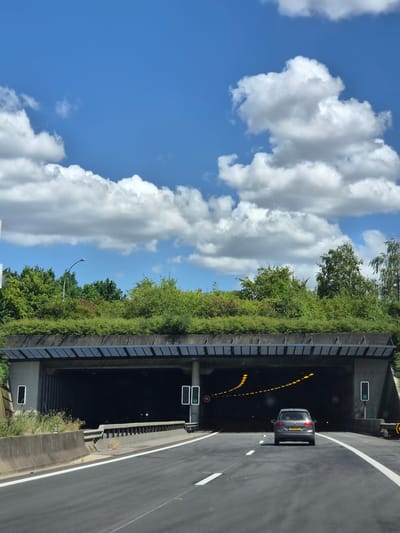The image shows a highway approach to a tunnel. The tunnel entrance is dark, with yellow lights lining the ceiling to guide vehicles. A silver car is visible entering the tunnel. Above the tunnel, the area is covered in lush green vegetation, with trees and bushes growing on the sloped sides and top. The sky is blue with large, fluffy white clouds, suggesting it is a bright, sunny day. The road has white lane markings and a concrete barrier on the right. Traffic signs are visible, including a green traffic light and speed limit signs indicating "50". The overall scene depicts a highway environment in a well-maintained, possibly European, setting, likely during daylight hours under clear weather. The location is identified as Frisange, Luxembourg. There is no visible text other than the numerals on the speed limit signs.