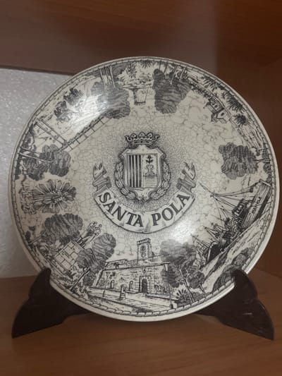 A decorative plate, likely a souvenir, is displayed on a dark wooden stand against a blurred background. The plate features a black and white illustration with a distressed, cracked glaze. The central design is a coat of arms with a crown on top, surrounded by a laurel wreath and a banner that reads "SANTA POLA". The illustration depicts a coastal scene with a fort or castle, trees, and sailing ships. The text suggests the location is Santa Pola, Spain. The overall scene is static and focused on the decorative object.