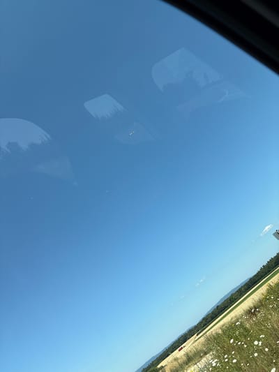 A wide, clear blue sky dominates the upper portion of the image. Reflections of trees and possibly a vehicle are visible on the windshield of a car. In the lower portion, a rural landscape unfolds, with a field of golden crops and a strip of white wildflowers bordering a dirt road. A red tractor is visible in the distance within the field. Hills covered in dark green trees form a backdrop to the field, and a few faint clouds are scattered across the sky. The image appears to be taken from inside a vehicle, looking out through the windshield. The scene is set in Ober-Olm, Germany.
