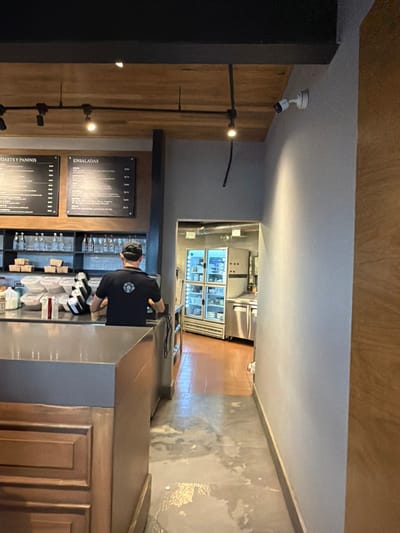 This image captures the interior of a commercial food establishment located in Fraccionamiento Los Heroes Queretaro, Mexico. The perspective is directed towards a service counter area and an open doorway leading to a rear kitchen or prep area.
In the foreground, a male individual is standing with his back to the camera, positioned behind a dark grey countertop. He wears a black short-sleeved uniform shirt featuring a white logo on the back and a black baseball cap. His hands are engaged over the counter.
Behind the individual, two rectangular black menu boards with white text are mounted on a light grey wall; visible titles include "TOASTS Y PANINIS" and "ENSALADAS." Below the menus, a dark shelving unit holds numerous clear bottles and white plastic containers. The service counter itself has a dark grey top, with a wooden paneled base and a metallic grey upper section. The immediate floor in the foreground is light grey concrete with irregular white splatter marks.
The ceiling above the counter is constructed of wooden slats, fitted with a black track lighting system containing multiple spotlights. A white dome-style security camera is mounted on the light grey wall in the upper right portion of the image, angled towards the front. A black cable segment hangs from the ceiling/wall just below the track lighting.
Through the open doorway, a brightly lit back area is visible, revealing commercial kitchen equipment. This includes at least two tall, multi-door refrigerators with glass fronts, stocked with various food items in containers. Several stainless steel surfaces, likely counters or appliances, are also present. The floor in this rear section is a reddish-brown tile. Metallic ventilation ductwork is partially visible near the ceiling of the back area.