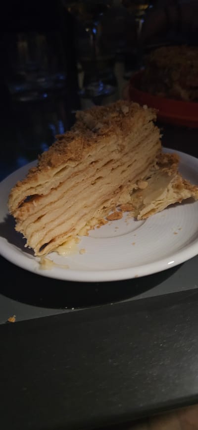 A slice of layered cake, likely a Napoleon or Mille-feuille, is presented on a white plate. The cake features numerous thin, flaky pastry layers interspersed with a creamy filling. The top and sides are dusted with crumbled pastry. The cake is positioned on a dark surface, with a blurred background suggesting an indoor setting. The overall lighting is dim, with some reflections visible in the background, hinting at glass objects and possibly a dimly lit restaurant or home environment. The context of Hévíz, Hungary, is not evident from the visual information provided in the image.