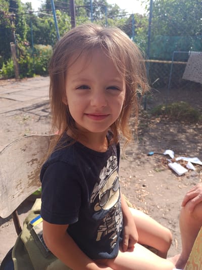 A young girl with long, light brown hair and fair skin is sitting on a wooden chair outdoors. She is looking at the camera with a slight smile, her eyes are dark and her eyebrows are light. She is wearing a dark blue t-shirt with a graphic print on it. Her arms are resting on her lap, and her legs are visible. Behind her, there is a dirt ground with some scattered leaves and twigs. In the background, there is a chain-link fence, some greenery, and a white cloth hanging on the fence. The scene appears to be in a rural or suburban setting, possibly a backyard or a courtyard. The lighting suggests it is daytime with sunlight. On the chair next to the girl, there is a green bag with a patch that resembles the Ukrainian flag. The overall mood of the image is casual and relaxed.