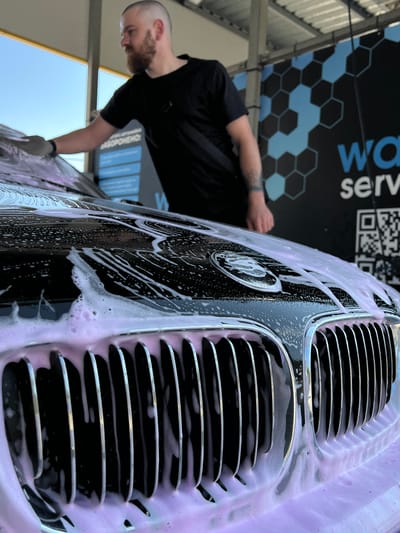 A man with a beard and a shaved head, wearing a black t-shirt, is washing a black car at an outdoor car wash. He is wearing a light-colored glove on his left hand and is in the process of foaming the car's hood and grill with a pink cleaning product. The car is a luxury model, with a prominent chrome grill and a BMW logo visible on the hood. Behind the car, a dark blue banner with a honeycomb pattern and white lettering, likely advertising the car wash service, is visible. The car wash appears to be a self-service facility, with a covered roof structure overhead. The scene suggests a bright, clear day, with the sky visible in the background. The location is identified as Velyki Hai, Ukraine.