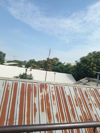 The image shows a view of rooftops and trees under a bright sky with scattered clouds. In the foreground, a rusty corrugated metal roof is visible, with streaks of orange-brown rust against a lighter gray or silver background. Beyond this, a white corrugated metal roof stretches across the middle ground, its ribbed texture clearly defined. Several trees with green and yellow foliage are scattered throughout the scene, with some appearing taller and more mature than others. In the distance, more rooftops and buildings can be seen, along with a construction crane. A tall antenna with multiple horizontal elements stands prominently on the right side of the frame. The overall impression is of a somewhat urban or semi-urban environment, possibly residential, bathed in daylight. No people are visible in the image. The scene is likely from a city with a temperate climate, given the foliage. Without specific landmarks, pinpointing the exact location within Osh, Kyrgyzstan, is not possible based solely on the visual information.