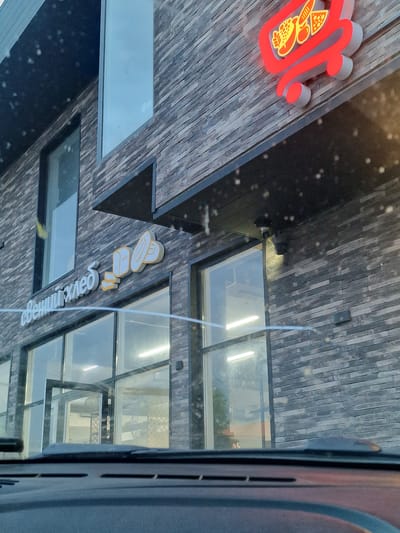 The image shows the exterior of a building with a brick facade. There are large windows and a dark overhang. A neon sign in the shape of a shopping cart with food items is visible on the upper part of the building. Below this, a sign in white letters reads "свєний хлєб" (Svein Khleb), which translates to "Svein's Bread". There are also stylized bread loaves depicted as part of the signage. Two security cameras are mounted under the overhang. The scene appears to be a storefront.