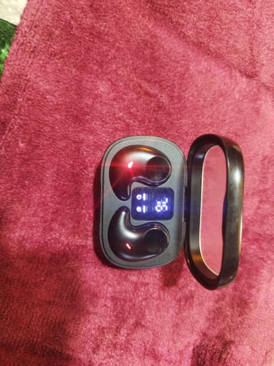 The image shows a pair of black wireless earbuds in their charging case. The case is open, revealing two earbuds nestled within their charging slots. A small digital display on the case shows "01" and "25" in blue LED lights, suggesting battery status or some other indicator. Red lights are visible on the earbuds themselves, likely indicating they are charging or powered on. The charging case is placed on a textured, deep red surface, possibly a rug or blanket. The background is out of focus but appears to be a similar red textured material. The scene is indoors. There are no people or other discernible objects in the image. The image does not contain any visual information that would suggest its location in Kyzyl-Kyya, Kyrgyzstan.