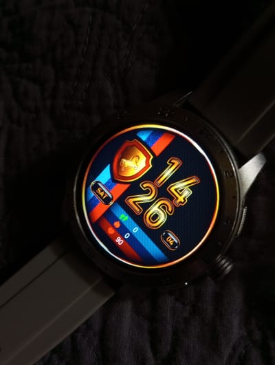 A close-up, overhead shot shows a smartwatch with a vibrant watch face displaying a digital clock, day of the week, and various health metrics. The watch face has a dark background with stylized stripes of red, blue, and orange. The time is displayed as "1:26", with the "1" being larger and positioned to the left of the "26". A shield emblem with a golden horse silhouette is visible at the top of the watch face. Below the time, there are icons for a heart with "90" next to it, and two green leaf icons with "0" next to them. A small oval with "04" is present on the right side of the watch face. The word "SAT" is displayed in an oval button to the left. The watch has a gray strap and is placed on a dark, textured fabric surface, possibly a tablecloth or blanket. The lighting is dim, creating a spotlight effect on the smartwatch. There is no information within the image to indicate the location in the city of Dymer, Ukraine.
