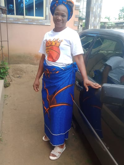 A middle-aged woman stands smiling gently at the camera, posing outdoors in Obehie, Nigeria. She is wearing a white short-sleeved t-shirt with a graphic featuring a basketball adorned with a golden crown. Her lower body is covered by a vibrant blue wrapper (skirt) with bold orange, abstract patterns and circular motifs. The edge of the wrapper displays visible text that reads "VERITABLE PRINTED BY". She also wears a matching blue and orange patterned headwrap or gele. On her feet are light-colored, embellished sandals with white soles.
Her right hand rests on the side mirror and door handle of a dark-colored car, possibly a sedan, parked beside her. A reflection of a person in a light grey t-shirt is visible in the car's window. In the background, to her left, is a light brown building with a window featuring blue-tinted glass panes. The ground is sandy or dirt, and a small amount of green foliage is visible near the building. The scene is brightly lit, suggesting it is daytime. The woman's expression conveys a pleasant and calm demeanor.