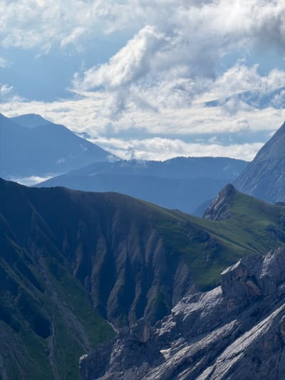 The image depicts a layered mountain landscape under a cloudy sky. In the foreground, rugged, dark grey rock formations dominate the lower right portion of the frame, showing significant texture and stratification. To the left and slightly behind these rocks, a steep, dark green mountainside descends, characterized by deep ravines and visible soil erosion. Further back, a prominent, rounded mountain ridge, also covered in dark green vegetation, creates a distinct curve across the middle ground. Beyond this ridge, multiple ranges of mountains recede into the distance, fading into progressively lighter shades of blue due to atmospheric perspective. The furthest peaks are shrouded in a light blue haze, and patches of white clouds are visible in the sky, some appearing dense and others wispy. The overall scene is a natural, unpopulated mountainous environment. This landscape is consistent with the topography of Wildermieming, Austria, a municipality known for its alpine setting.