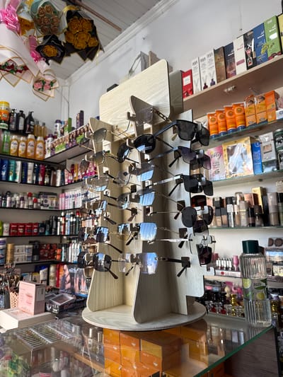 The image depicts the interior of a retail establishment, likely a cosmetics or accessories shop, situated in Yovon, Tajikistan. The scene is filled with various products displayed on shelves and a rotating stand. Prominently featured is a display rack holding numerous pairs of sunglasses, varying in style and color. Behind the sunglasses display, shelves are stocked with an assortment of cosmetic items, including bottles, tubes, and boxes. Some of these products appear to be shampoos, lotions, and fragrances, with labels indicating brands and scents. Further back, on higher shelves, more products are visible, including boxed items and what seem to be gift sets or perfumes. Decorative bouquets of artificial flowers are hanging from the ceiling in the upper left portion of the image. The overall environment suggests a commercial space with an emphasis on beauty and personal care products. No individuals are present in the frame, and the focus is solely on the merchandise and its arrangement.