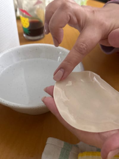 A close-up, overhead shot shows a person's hands holding a translucent, light-colored object above a white bowl filled with clear liquid. The object, which is flexible and appears to be made of silicone, has a rounded, almost tear-drop shape. The person's left hand, with light pink nail polish, holds the object from its upper edge, while the thumb and index finger of the right hand gently touch its lower edge, poised above the water in the bowl. This suggests the object is being prepared for washing, rinsing, or placement into the liquid.

The white ceramic bowl features small dark speckles on its interior surface and rim. In the lower-left corner, part of a clear glass bottle with a yellow pineapple design and red triangular motifs is visible. To the right, a folded kitchen towel with green and white stripes lies on a wooden surface, which also forms the background for the scene. The lighting indicates an indoor setting, likely a domestic kitchen or dining area. There is no visible text in the image.
