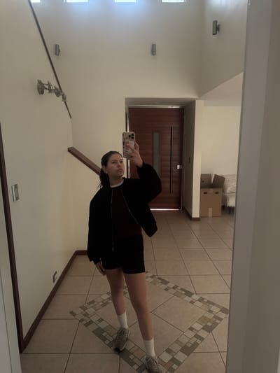 A mirror captures a woman in an indoor setting in San Pedro Cholula, Mexico. The woman holds a phone to take a picture of herself wearing a black jacket, shorts, and white socks with sneakers. The reflection shows a hallway with beige tiled floors featuring a decorative border pattern. A dark brown door is visible at the end of the hall, with boxes on a shelf above the door. The wall has light fixtures and a decorative coat rack. Daylight enters through a window.