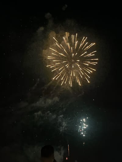 The image depicts a nighttime scene with fireworks. A large, golden firework is exploding in the upper center of the frame, its radiant streaks filling a significant portion of the dark sky. Below and to the right, a smaller cluster of white and yellow fireworks is visible, some appearing as bright, distinct orbs. Faint wisps of smoke from previous fireworks are scattered throughout the darkness. In the lower foreground, the dark silhouette of a person's head and shoulders is visible, looking upwards at the display. The scene is suggestive of a public fireworks event, possibly occurring during a festival or celebration in Castelló d'Empúries, Spain.