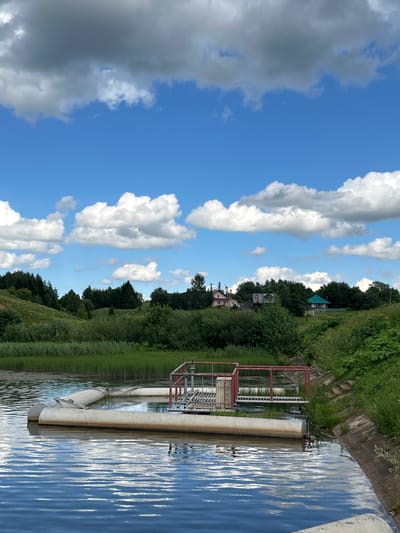 In the city of Verkh-Rozhdestvo, Russia, a body of water is visible, likely part of the settlement's water management system. In the foreground, large, white pipes float on the surface, partially submerged. Adjacent to these pipes is a metal grate structure with red railings, situated on a platform within the water. The water itself is calm, reflecting the blue sky and white clouds that dominate the upper portion of the image. Beyond the water, a bank of tall green grass and reeds lines the edge, leading to a gently sloping hill covered in lush green trees. In the distance, several houses are nestled amongst the trees, some with visible roofs of various colors, including pink and green. The overall scene depicts a rural landscape with elements of industrial infrastructure integrated into the natural environment.