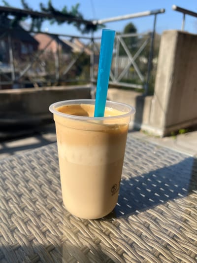 A clear plastic cup containing a layered iced beverage, approximately two-thirds full, rests on a light-colored, woven synthetic rattan table. The beverage consists of a lighter, opaque lower layer and a darker, frothy upper layer. A light blue, translucent cylindrical straw is inserted into the drink. Strong, direct sunlight illuminates the scene, casting distinct shadows from the cup and the table's weave onto the table surface.
In the background, out of focus, a metal railing system with horizontal bars and vertical supports is visible, partially obscured on the left by green foliage. Beyond the railing, several residential buildings with reddish-brown pitched roofs are discernible under a clear blue sky. A concrete support pillar forms part of the railing structure on the right. The scene depicts an outdoor setting, likely a balcony or terrace, in the city of Würselen, Germany. No individuals or specific actions are present.