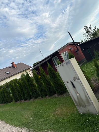 The image captures a residential scene in Jelgava, Latvia, under a partly cloudy sky. In the foreground, a gravel path borders a manicured lawn. A tall, grey electrical cabinet, bearing a yellow and black high-voltage warning symbol, stands prominently on the right side of the frame. Behind it, a row of evenly spaced, conical evergreen trees forms a lush green hedge.
Further back, two houses are visible. The house on the left has a light-colored facade, a dark brown tiled roof, and a red brick chimney. A small dormer window is present on the roof. To its right, a red wooden house with a dark roof features a prominent black metal chimney pipe extending upwards. A solar panel is affixed to the wall of the red house. A dark fence encloses the property. The overall impression is one of a quiet, suburban or semi-rural setting.