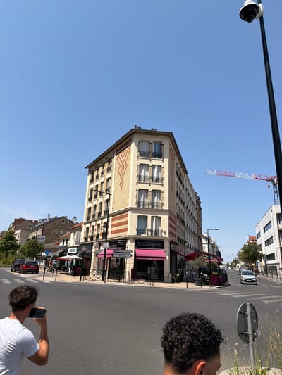 The image depicts a street scene in Courbevoie, France, on a clear, sunny day. In the foreground, the backs of two men are visible. The man on the left is wearing a white t-shirt and holding a smartphone to his ear, as if on a call. The man on the right has curly dark hair. The street is paved with asphalt and features a pedestrian crossing.

Dominating the mid-ground is a multi-story building with a distinctive facade featuring cream-colored bricks and red horizontal banding. The building has several windows with shutters. At street level, businesses are visible, including a "Boulangerie Pâtisserie" with a pink awning and outdoor seating. Signage indicates directions to "Centre Hospitalier Rives de Seine" and "Site de Courbevoie."

To the right of the main building, another contemporary white building stands. Further down the street, traffic is present, with a white car visible in the distance. A construction crane with red and white elements is visible in the sky to the right, suggesting ongoing development in the area. A lamppost with a surveillance camera is prominently positioned on the right side of the image. Lush green trees are interspersed along the street. The sky is a clear, vibrant blue.